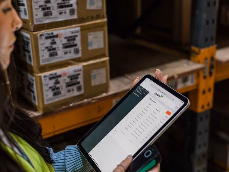 Worker holding a tablet and barcode scanner in a warehouse with stacked boxes.