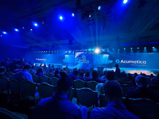 Audience seated in blue-lit conference room with Acumatica Summit logos on large screens and stage.