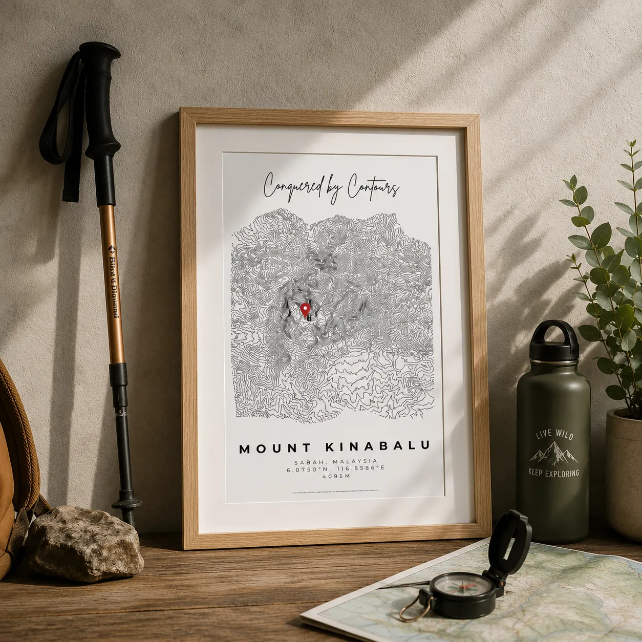 Framed contour map of Mount Kinabalu with trekking pole, compass on map, water bottle, and plant on table.