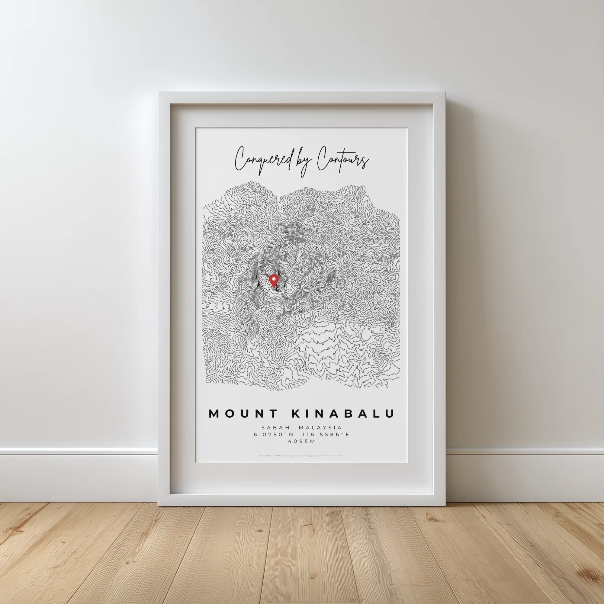 Framed contour map of Mount Kinabalu with a red location marker on a wooden floor against a white wall.