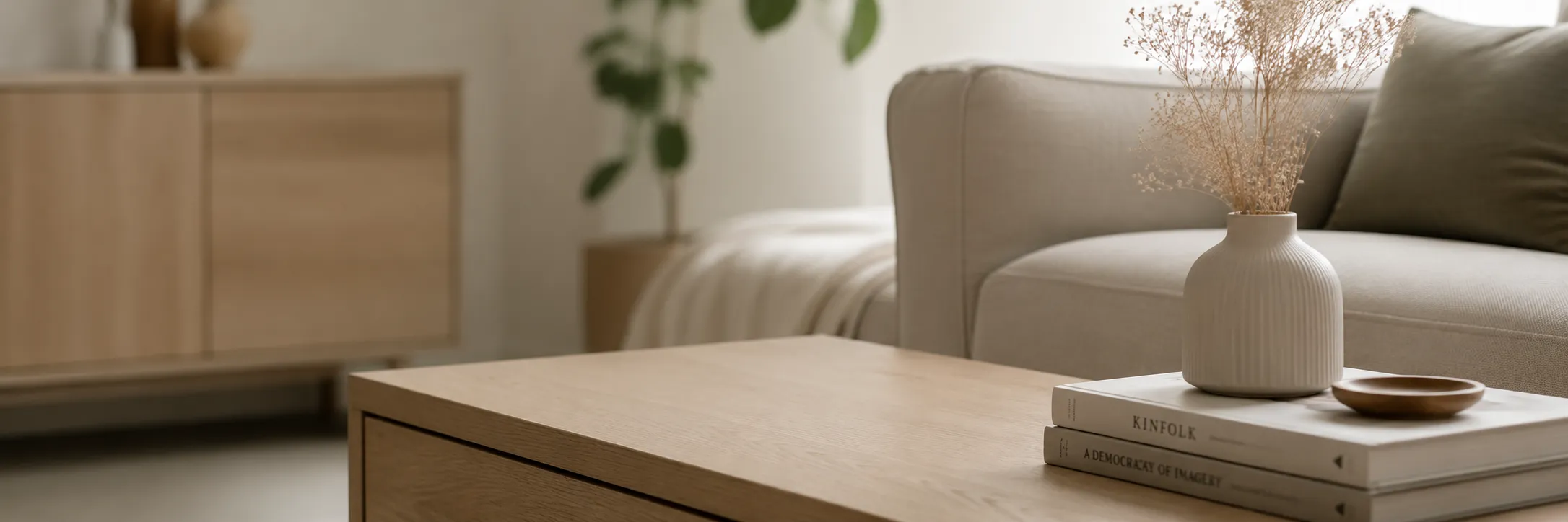 Light wooden coffee table with stacked books, a vase with dried flowers, beige sofa, and blurred background.