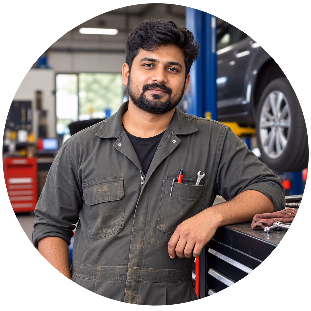 A mechanic in grey overalls leaning on a tool chest, a spanner in his pocket and a car on a ramp behind.