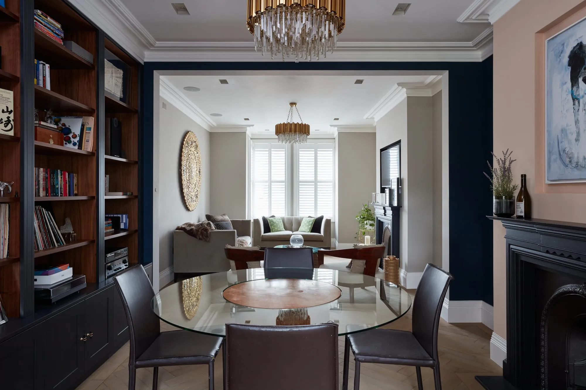 Dining room with a round glass table, navy walls and built-in shelving, opening onto a sitting room with shutters