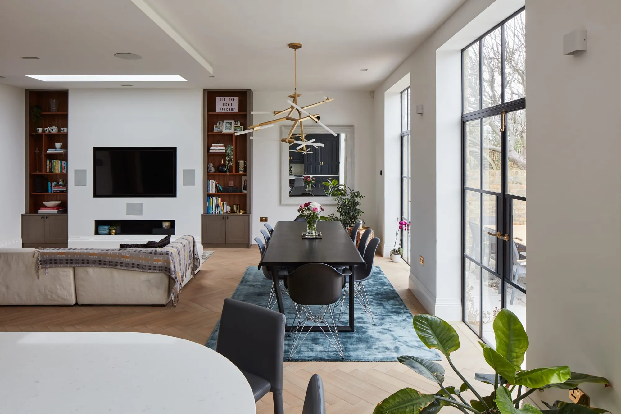 Open-plan living and dining room with tall steel-framed windows, a media wall and herringbone floor