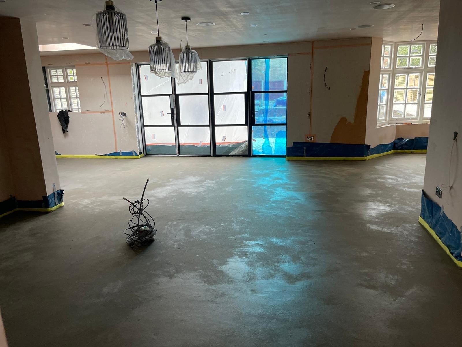 Empty open-plan ground floor with fresh screed, pendant lights and black-framed glazed doors