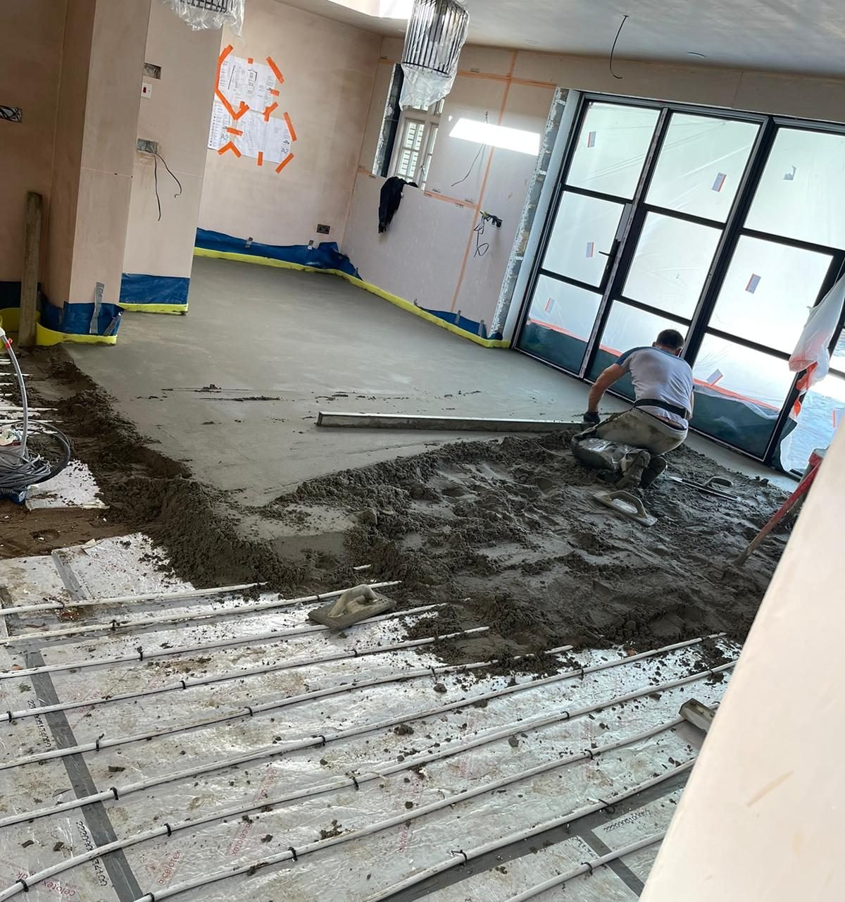 Builder spreading screed over underfloor heating pipes in front of the glazed doors