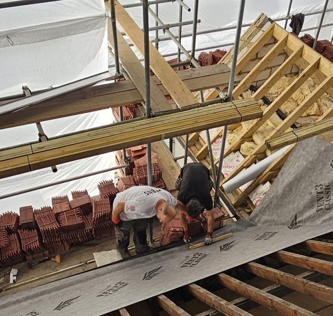 Two BNS roofers laying breathable membrane beside stacks of clay tiles, with exposed rafters above