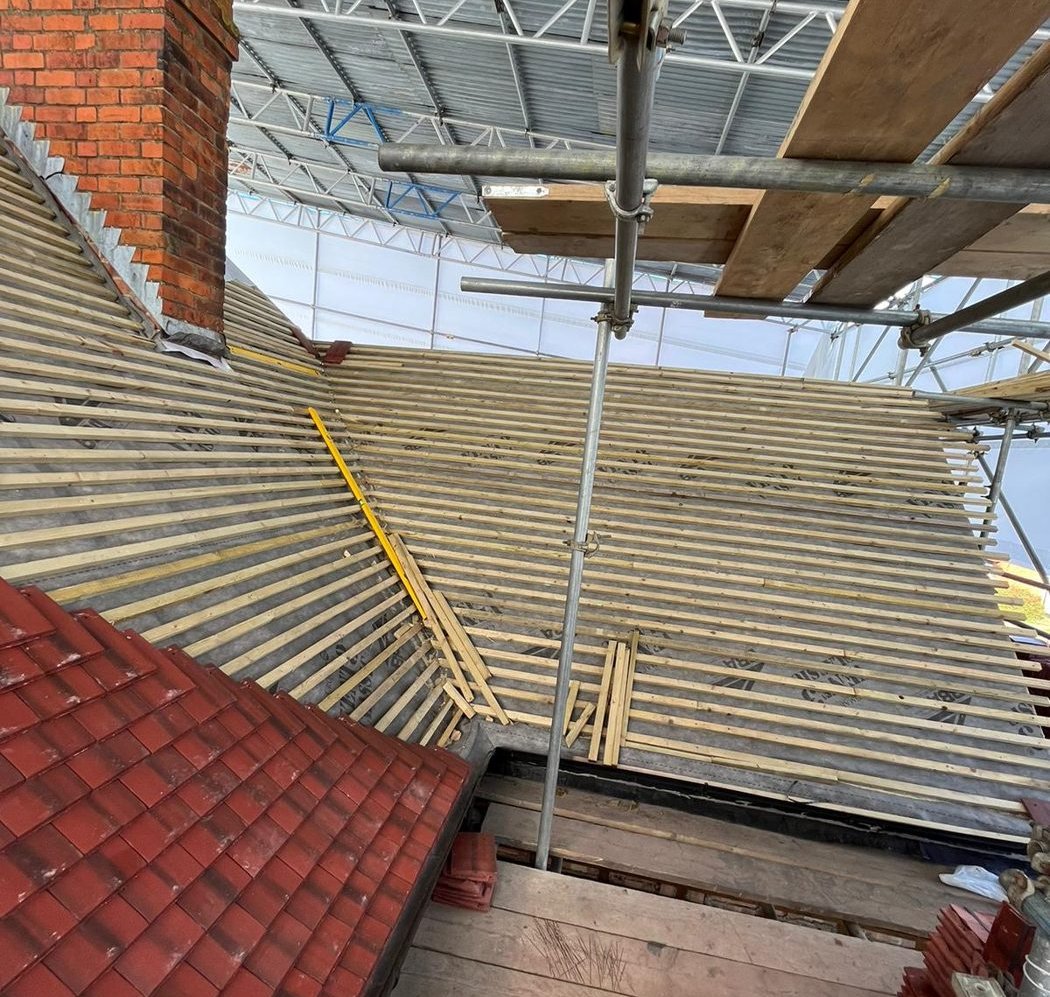 Roof slopes battened out under a temporary scaffold roof, next to a finished section of red tiles