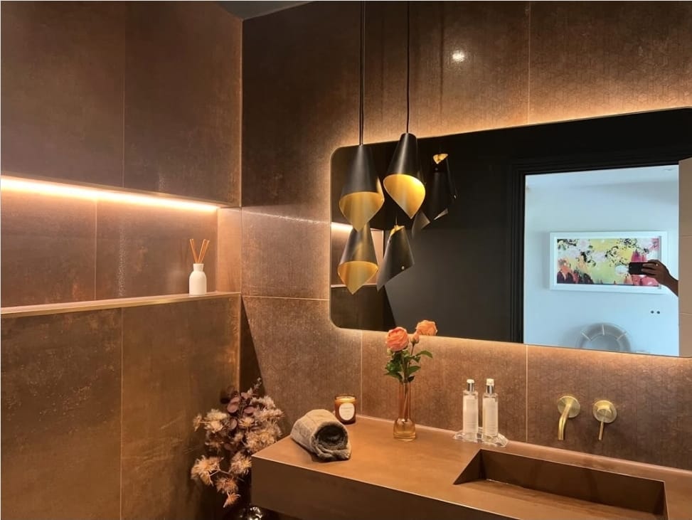 Cloakroom with bronze wall tiles, a backlit mirror, black and gold pendant lights and brass taps