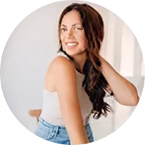 Smiling woman with long brown hair wearing a white tank top and jeans, posing indoors.
