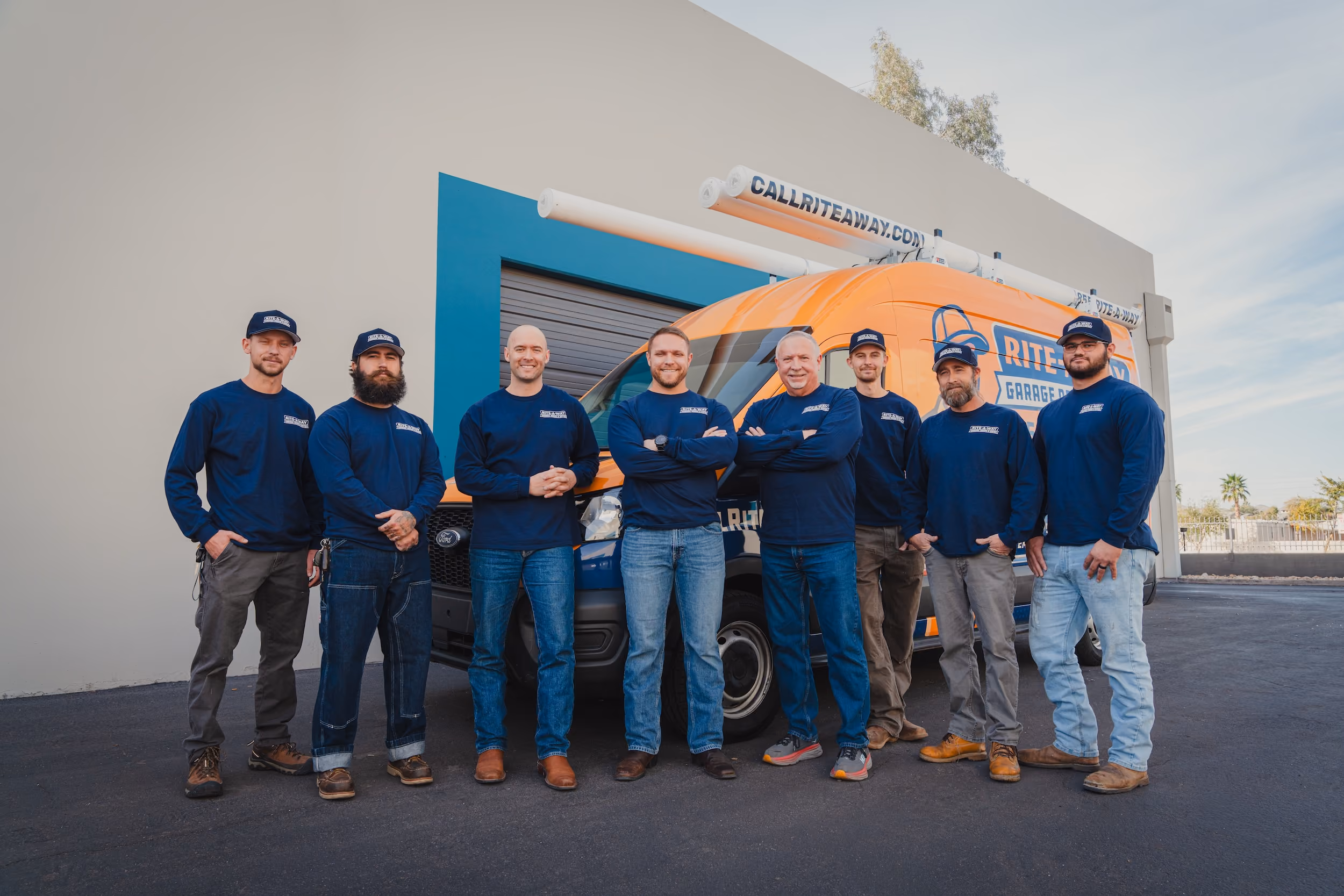 Eight men in matching blue shirts and hats standing in front of a branded orange and blue service van.