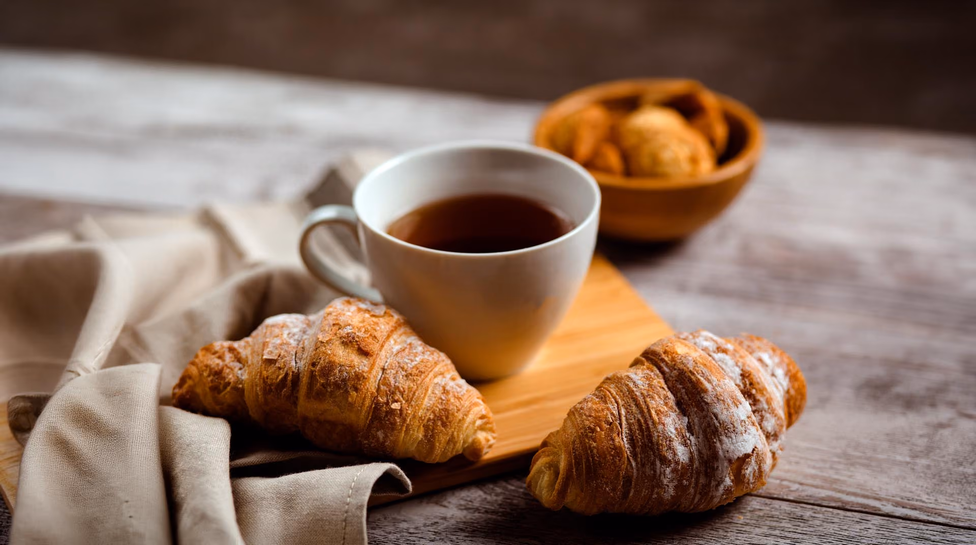 Hvite kopp med te mellom to croissanter på et trebord med en brun klut og bolle i bakgrunnen.