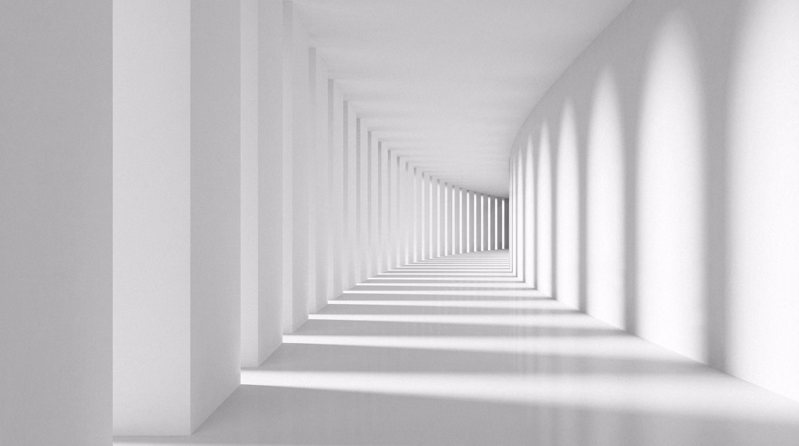 Un couloir blanc lumineux avec des colonnes alignées formant un motif d'ombres et de lumière répétées sur le sol et les murs.