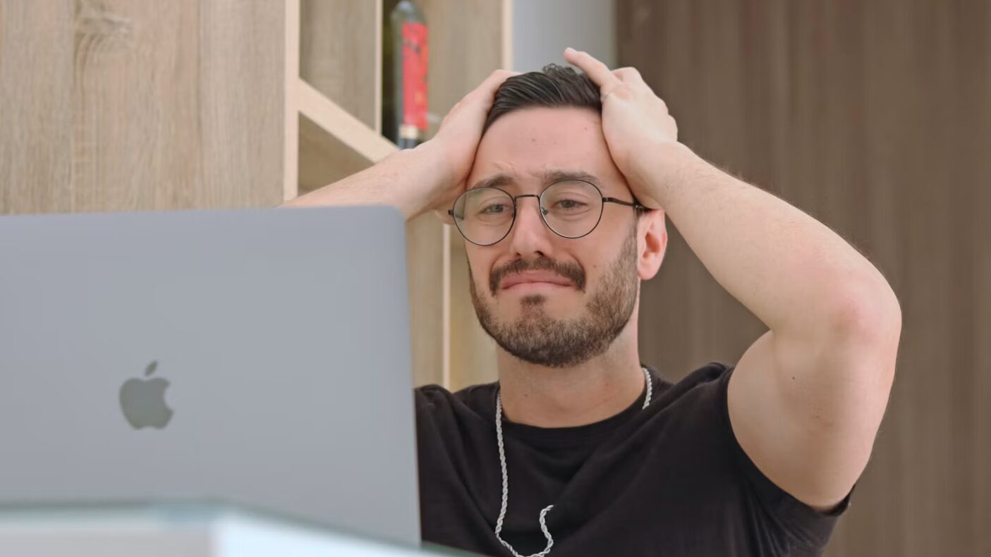Homme dépité, les mains sur la tête, devant son MacBook