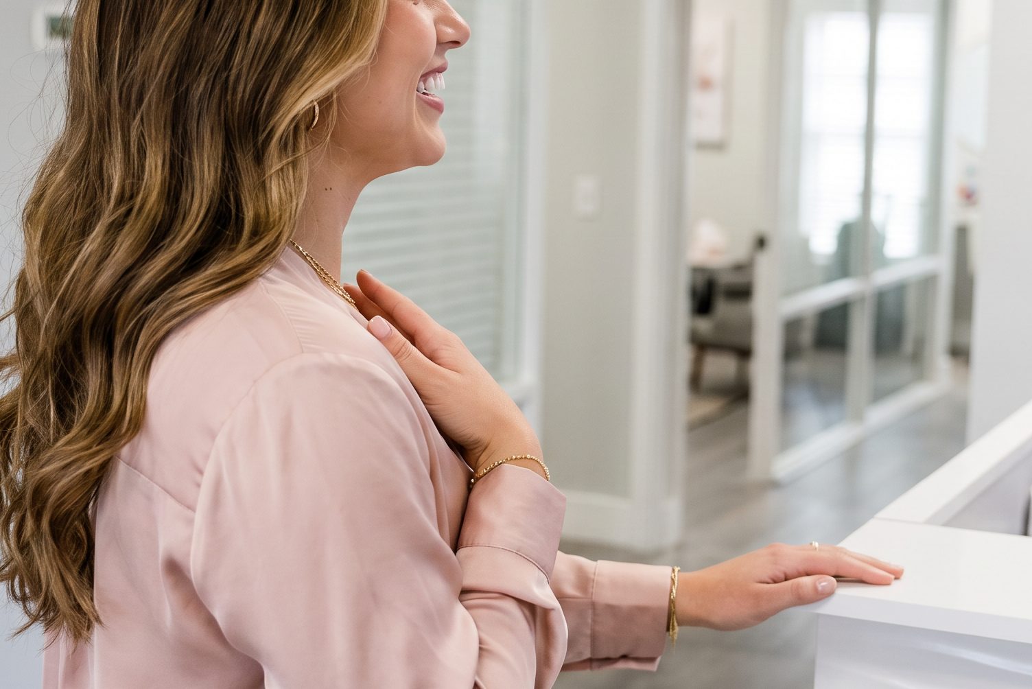 Smiling woman in a blush blouse resting her hand on the white wave-front reception counter