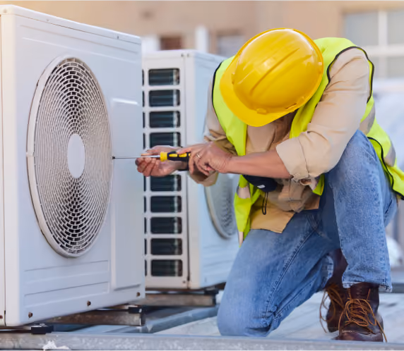 A worker wearing a yellow hard hat, safety vest, and jeans is kneeling and using a screwdriver to repair or install a large white air conditioning unit outdoors.