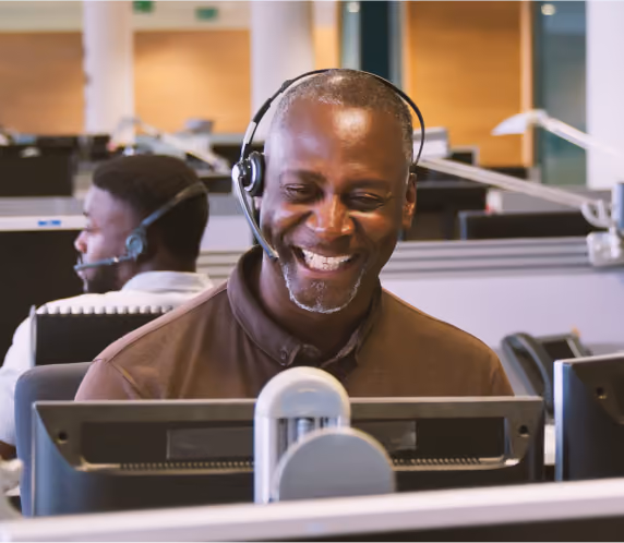 Smiling man wearing a headset working at a computer in a busy call center with another operator in the background.