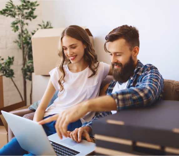 A smiling young couple sitting on a couch looking at a laptop together, surrounded by cardboard boxes and a potted plant, suggesting they are moving or unpacking.