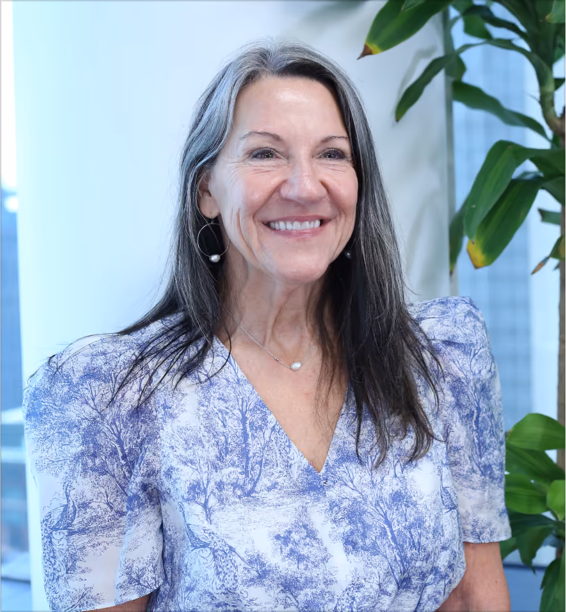 Smiling middle-aged woman with long gray and dark hair wearing a white and blue patterned blouse and hoop earrings, standing indoors near green plants.