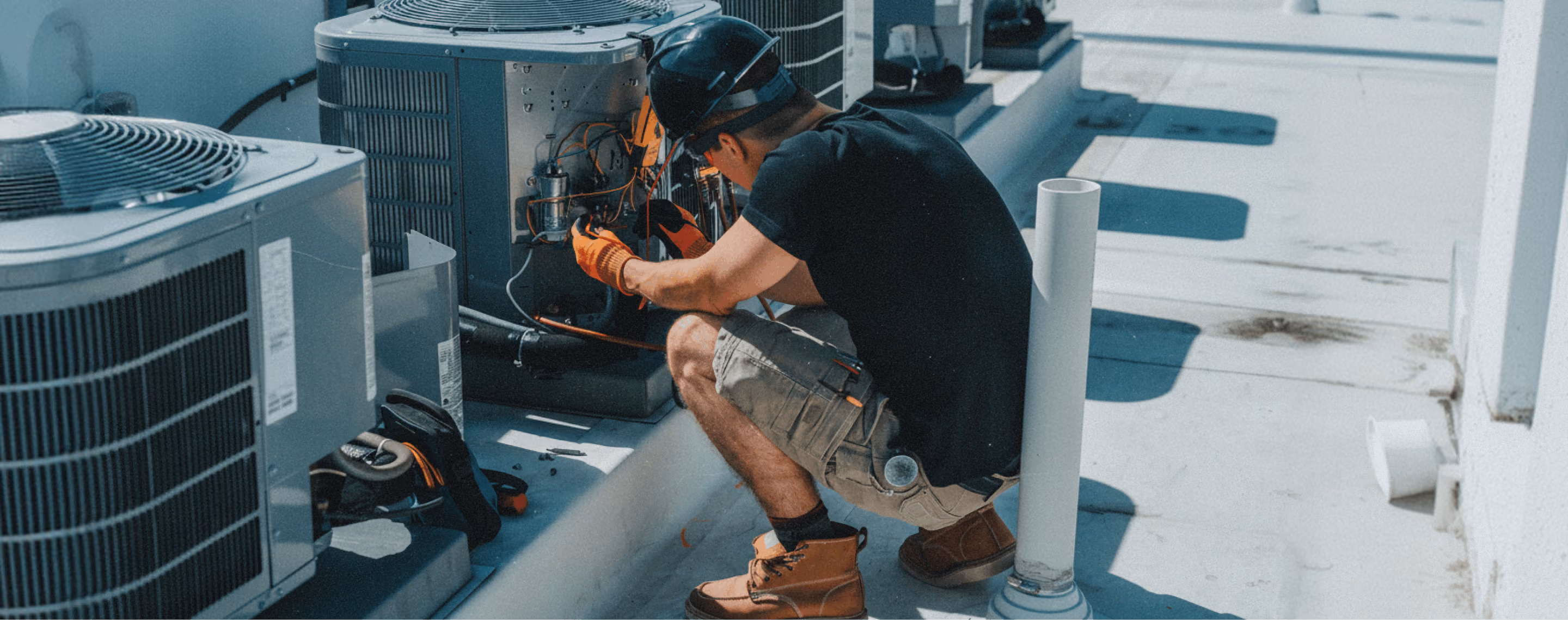 Technician wearing a helmet and gloves repairing an outdoor air conditioning unit on a rooftop during daytime.