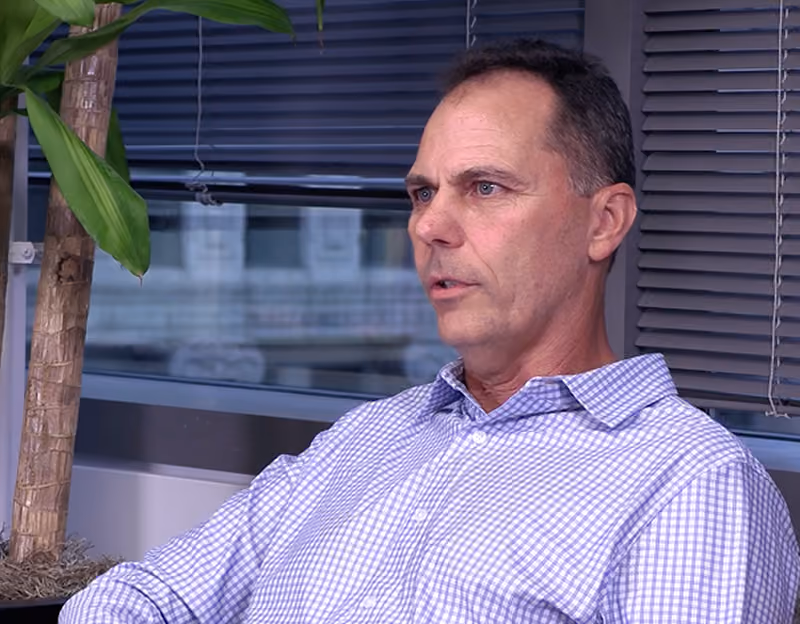Middle-aged man with short dark hair wearing a light purple checkered shirt sitting indoors near a window with closed blinds and a tall green plant.