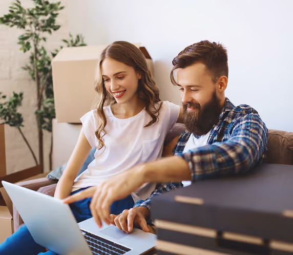 Smiling young couple sitting on a couch looking at a laptop together, surrounded by cardboard boxes and a plant in the background.