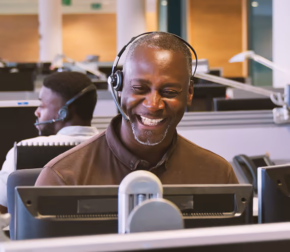 Smiling man wearing headset working at a computer in a call center with another person visible in the background.