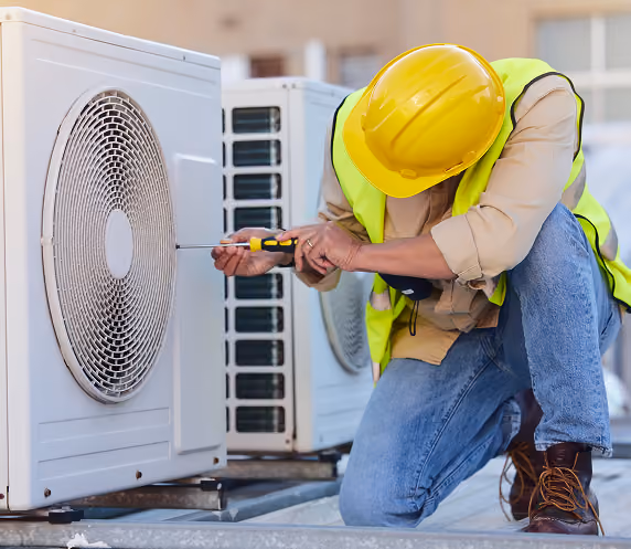 A worker wearing a yellow safety helmet and neon vest kneels on one knee while using a screwdriver to repair or maintain an outdoor air conditioning unit.
