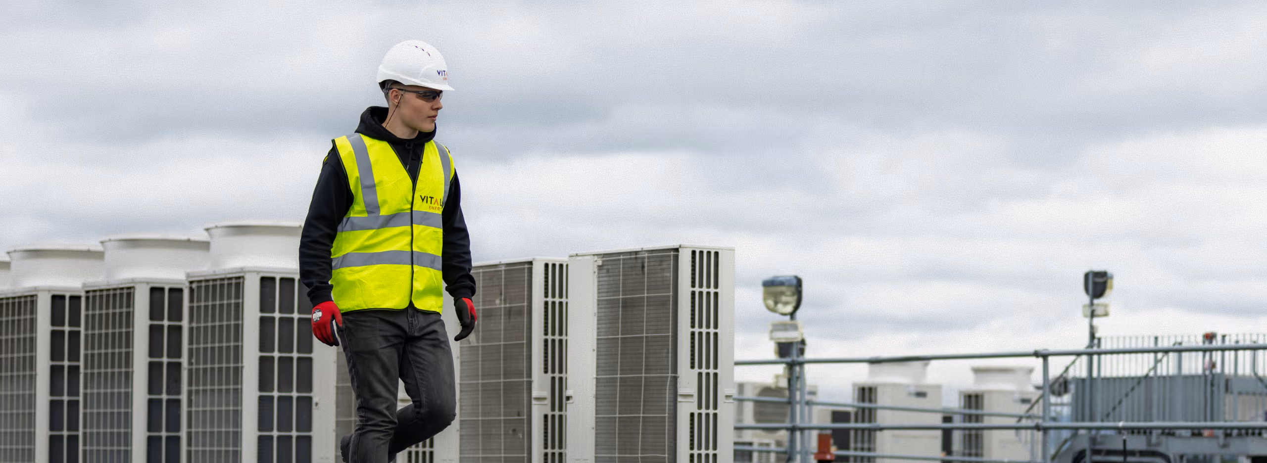Worker in safety helmet, gloves, and yellow reflective vest walking on rooftop near several large HVAC units under cloudy sky.