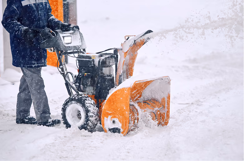 Person operating an orange snow blower clearing snow on a snowy day, with snow being thrown out to the side.
