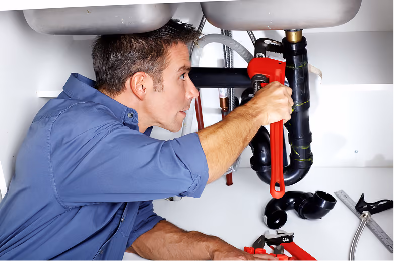 Man in a blue shirt using a red pipe wrench to fix plumbing under a sink, with various tools and pipe parts nearby.