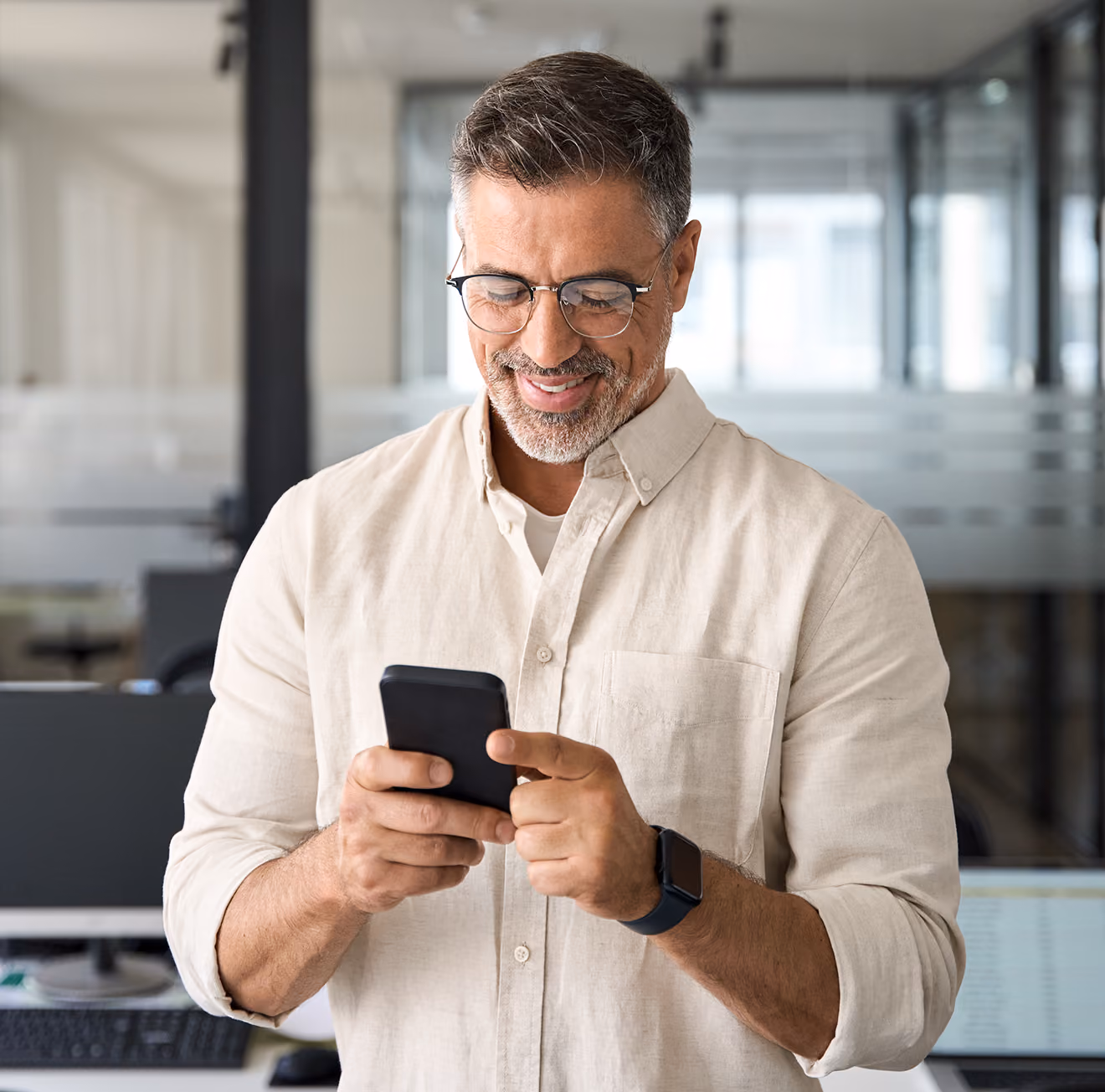Middle-aged man wearing glasses and a light beige shirt smiling while using a smartphone in a modern office setting with computer monitors and glass partitions in the background.