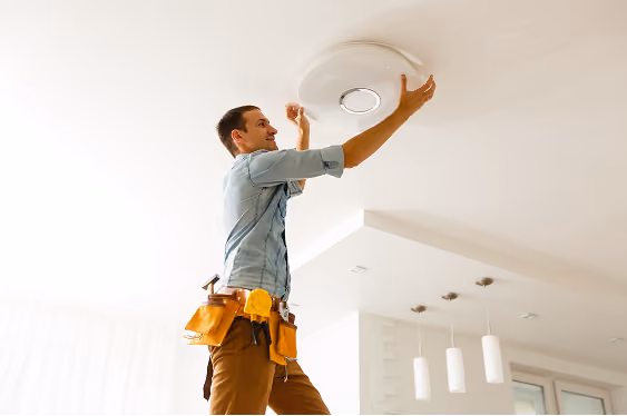 Handyman in a tool belt installing or repairing a ceiling light fixture in a modern, bright room.