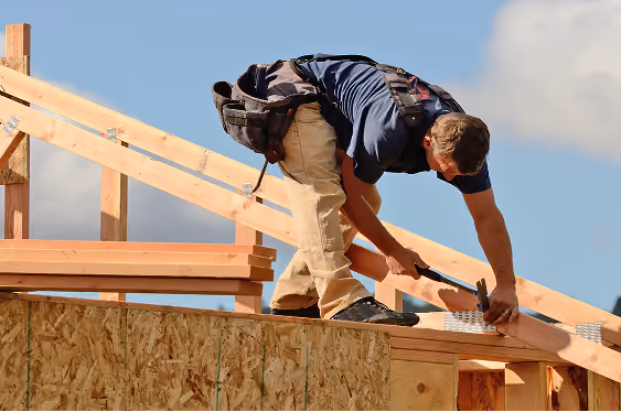 Construction worker wearing a tool belt nailing wooden beams on a house roof frame under a blue sky.
