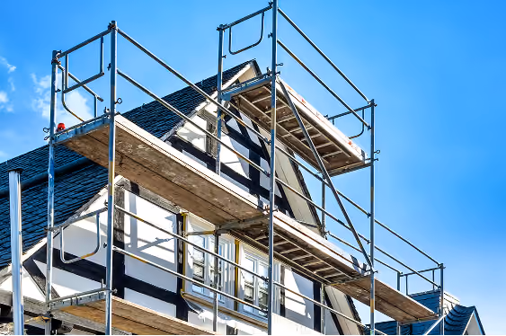 White house with dark roof surrounded by metal scaffolding against a clear blue sky, indicating construction or renovation work.