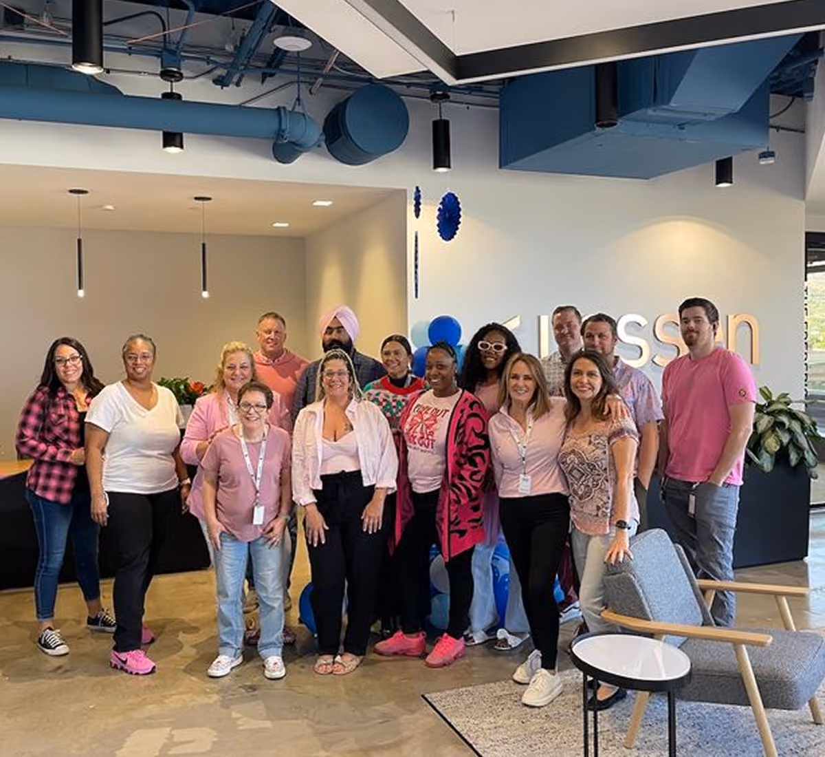 Group of fifteen diverse adults, mostly women, smiling and posing together in an office lobby with a light-colored floor, modern ceiling, and a 'Kission' sign partially visible on the wall behind them.