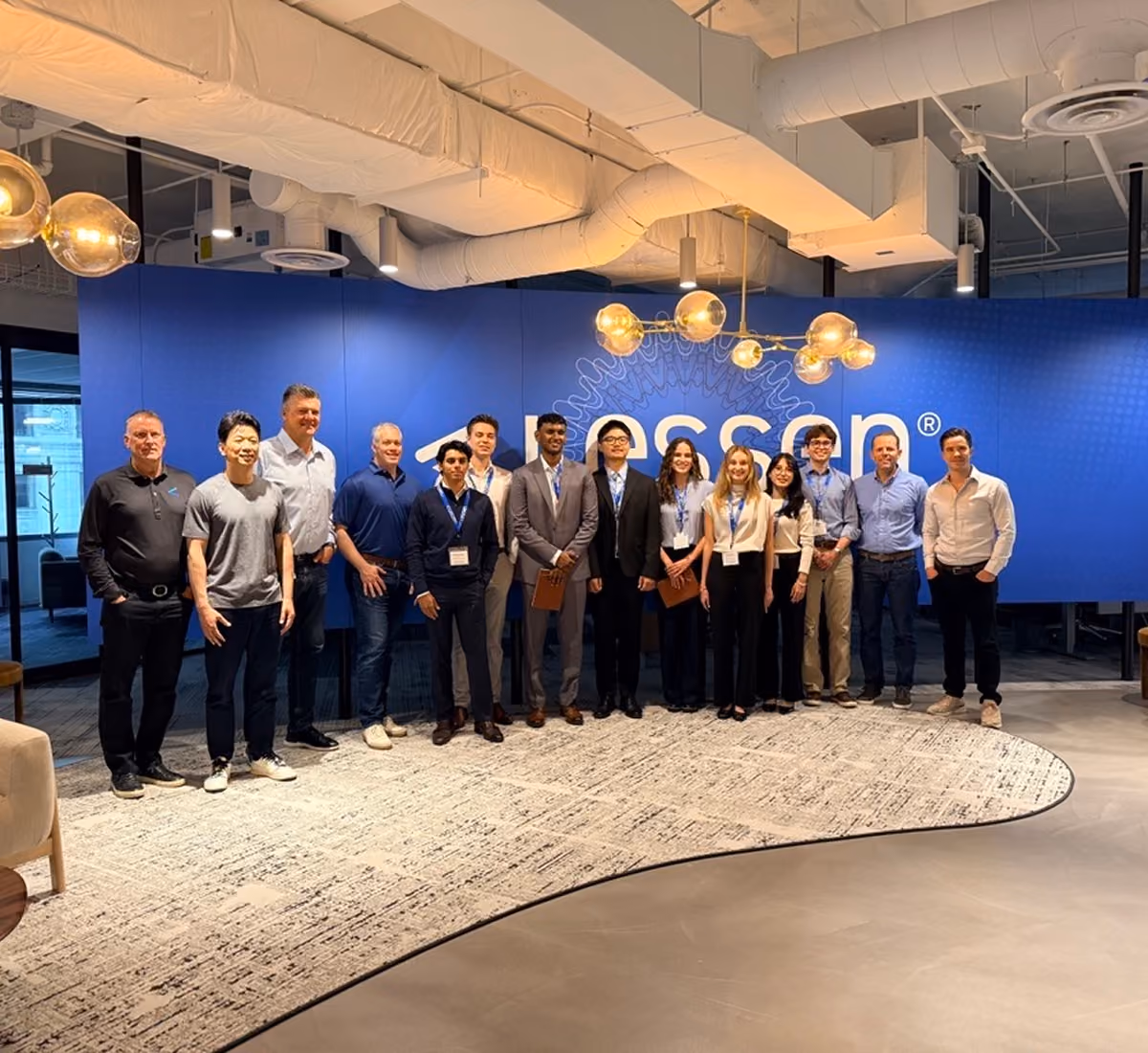 A group of fourteen diverse professionals standing side by side in a modern office space with a blue wall and a large logo behind them, under warm hanging lights.