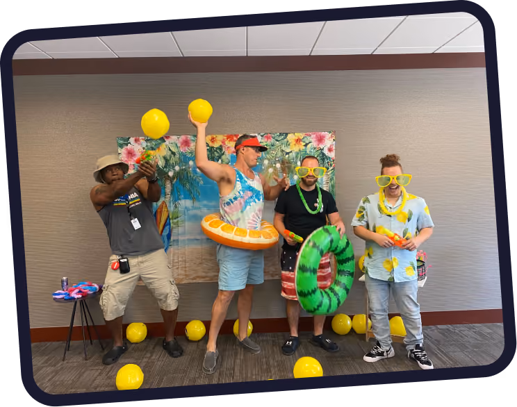 Four men posing in a playful summer-themed setting indoors with tropical background. One man on the left wears a bucket hat and holds yellow balloons, the second wears a visor and an orange inflatable ring holding more balloons, the third wears oversized yellow glasses and holds a green inflatable ring, and the fourth also wears oversized yellow glasses and a lemon-patterned shirt, holding a small orange object. Yellow balloons are scattered on the floor around them.