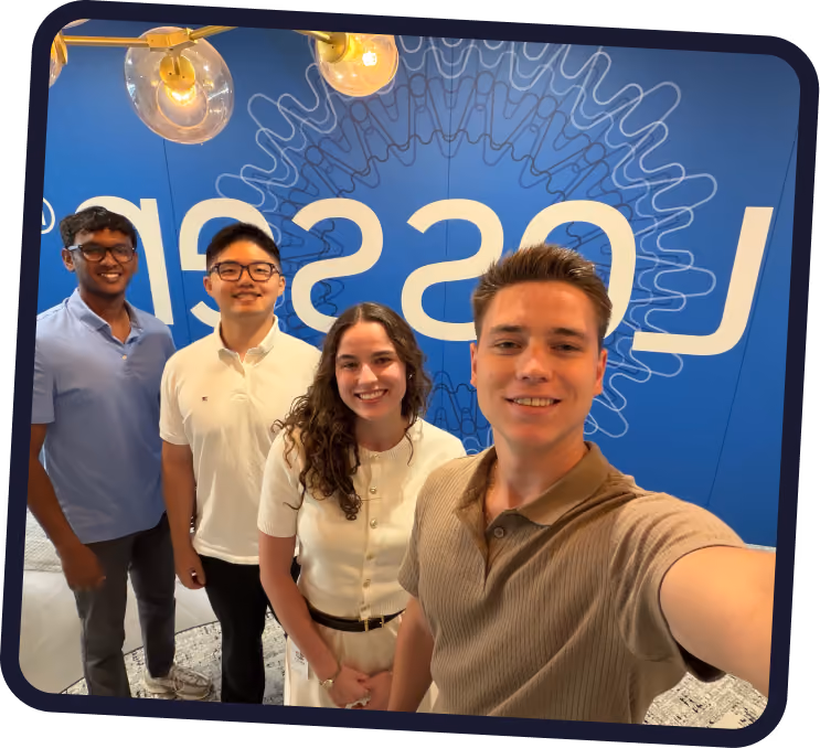 A group of four young adults smiling for a selfie in front of a blue wall with a large partially visible white logo and decorative line pattern.