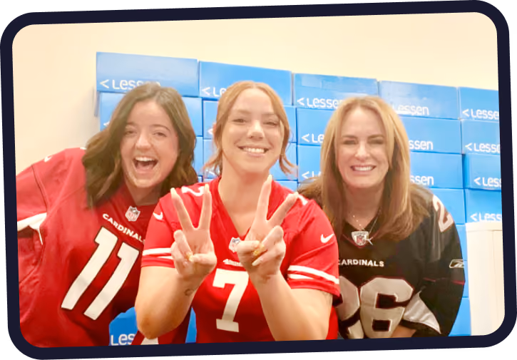 Three smiling women wearing Arizona Cardinals football jerseys stand in front of a wall stacked with blue boxes labeled 'Lessen'.
