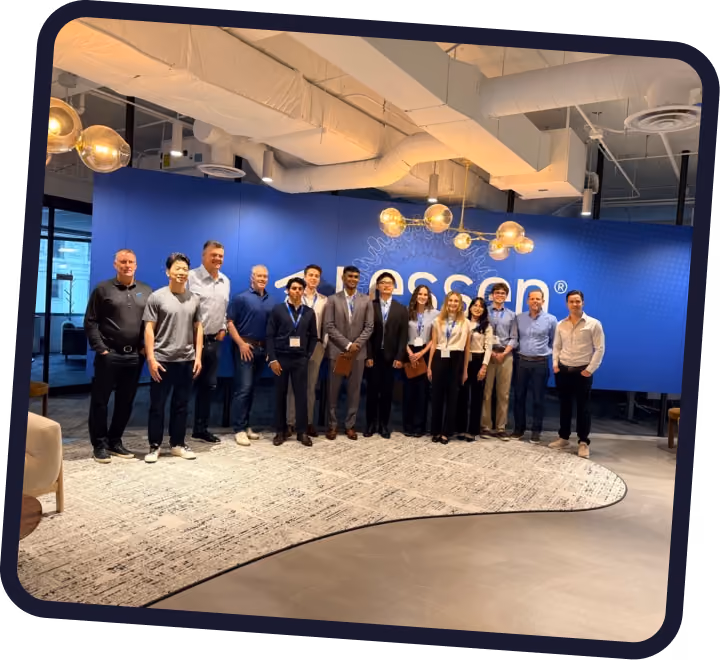 Group of thirteen diverse professionals standing side by side in a modern office with a blue wall behind them displaying the word 'Accera.' The office features hanging globe lights and exposed ceiling pipes.
