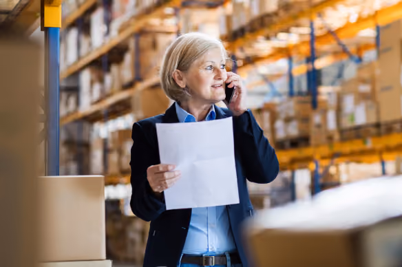 A woman in business attire standing in a warehouse, holding a paper in one hand and talking on a mobile phone, with shelves of boxes in the background.