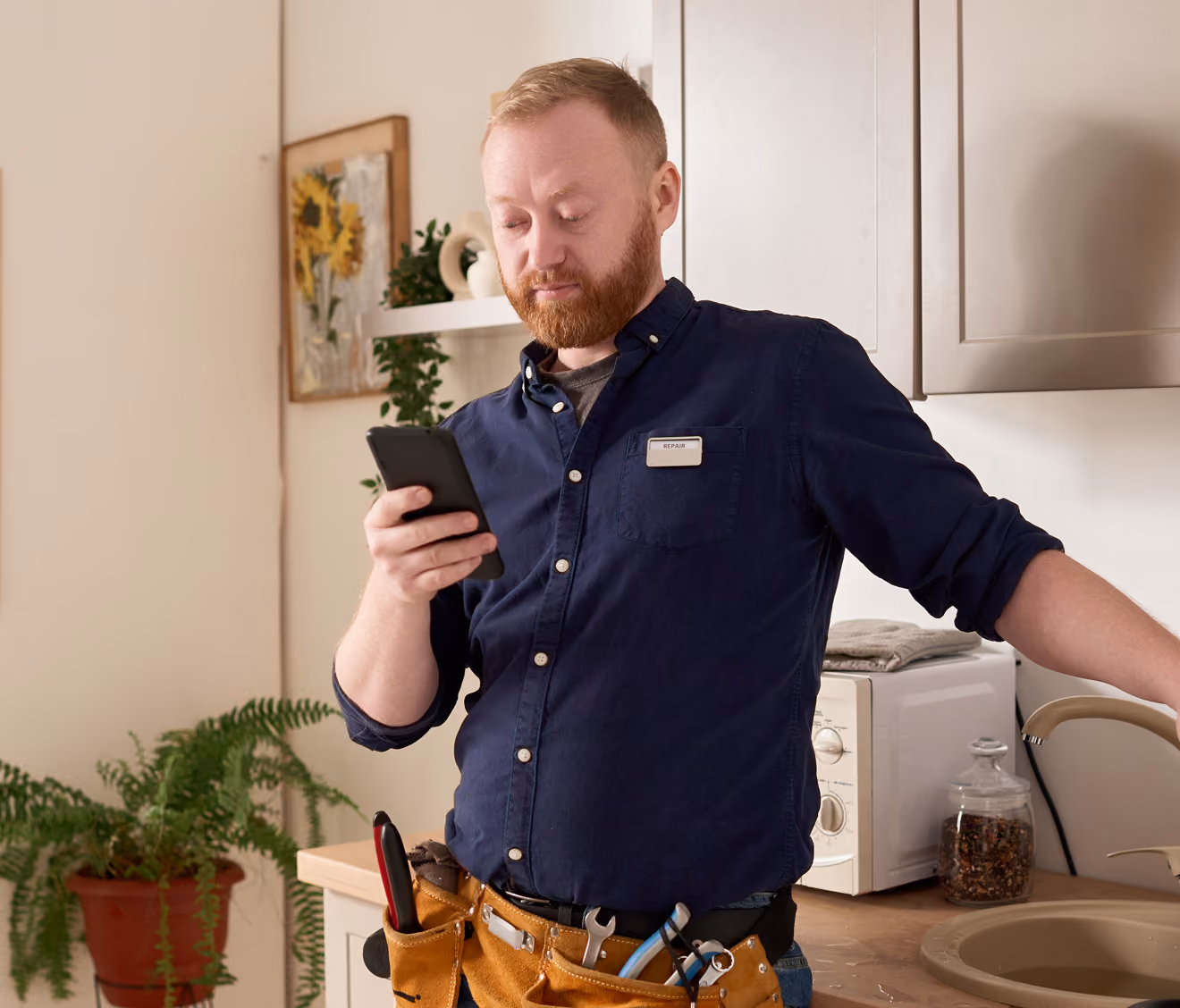 Man with a beard wearing a navy blue shirt and a tool belt looking at a smartphone while standing in a kitchen near a sink and microwave.