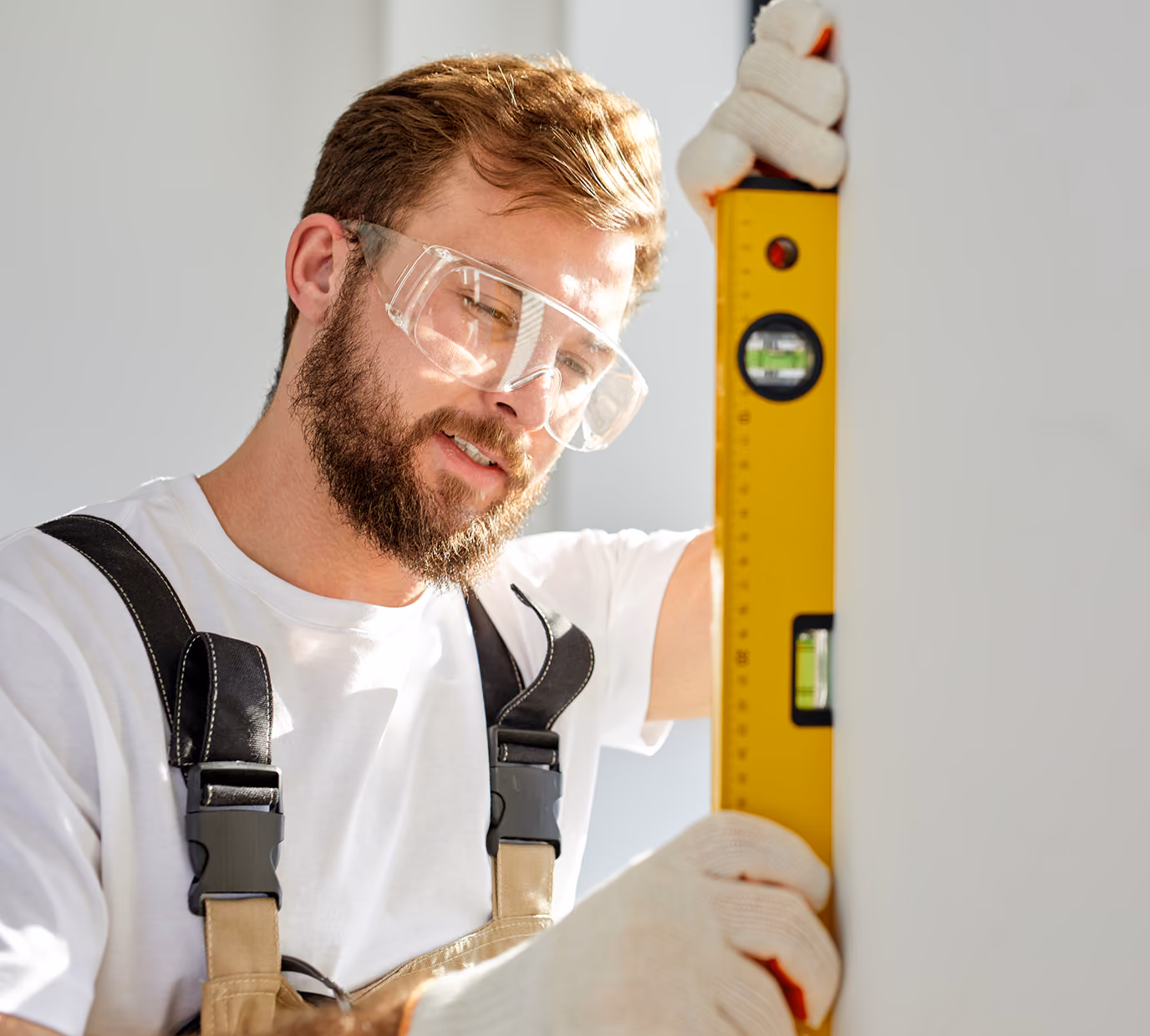 Bearded man wearing protective glasses and work gloves holds a yellow spirit level against a wall, checking for plumb.