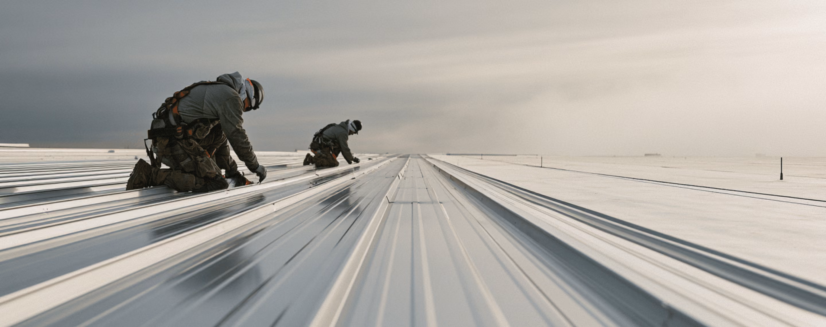 Two workers wearing safety gear and helmets kneeling on a large metallic roof while performing installation or maintenance against a cloudy sky background.