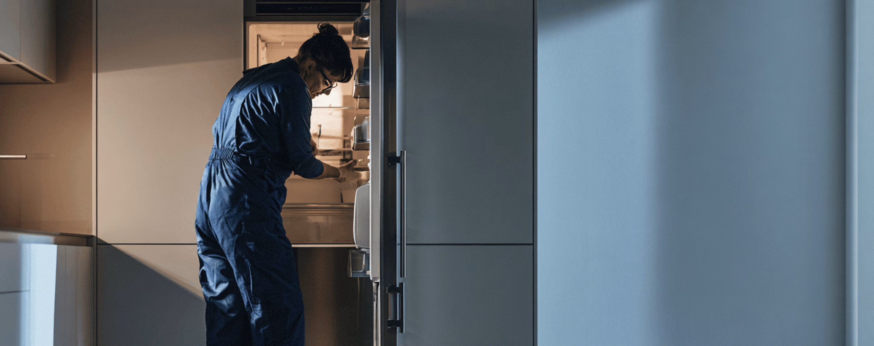 Person wearing a dark coverall opening and inspecting the inside of a refrigerator in a dimly lit modern kitchen.