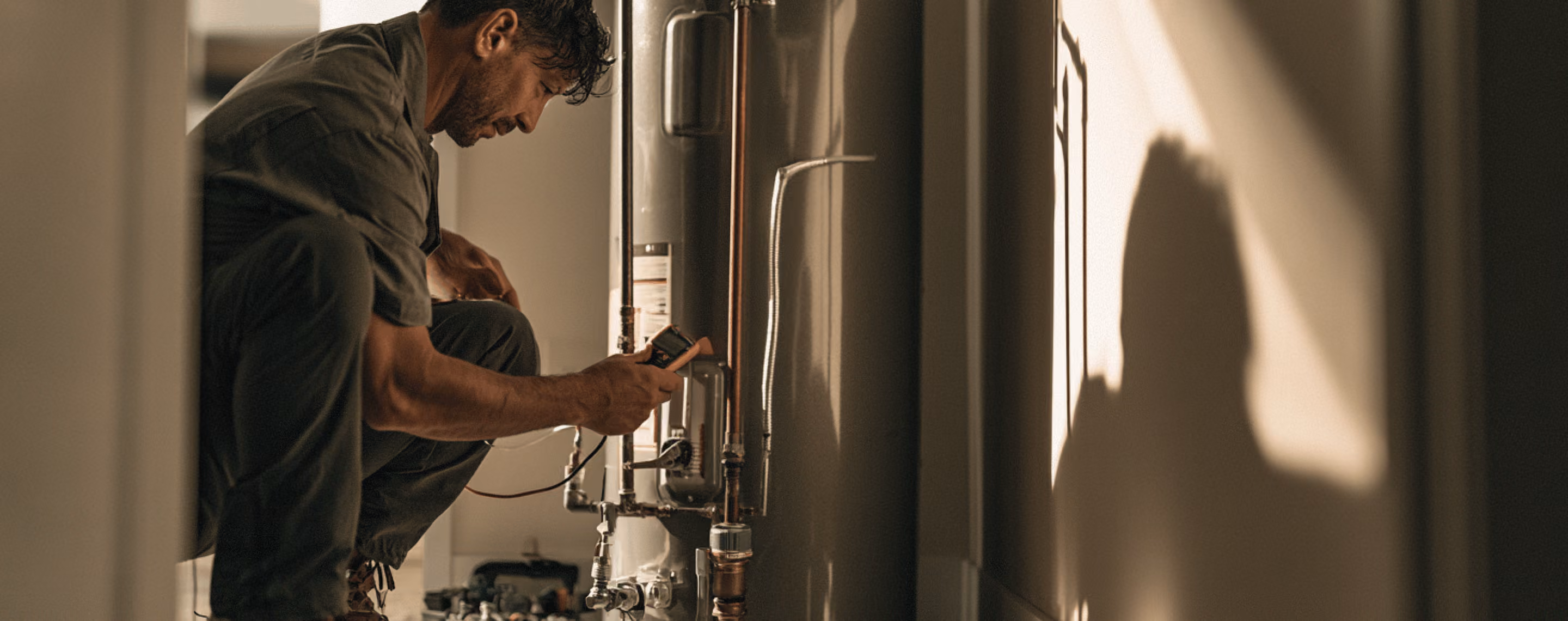 Man crouching and using a device to inspect or repair a metal water heater or boiler in a dimly lit room with a shadow cast on the wall.