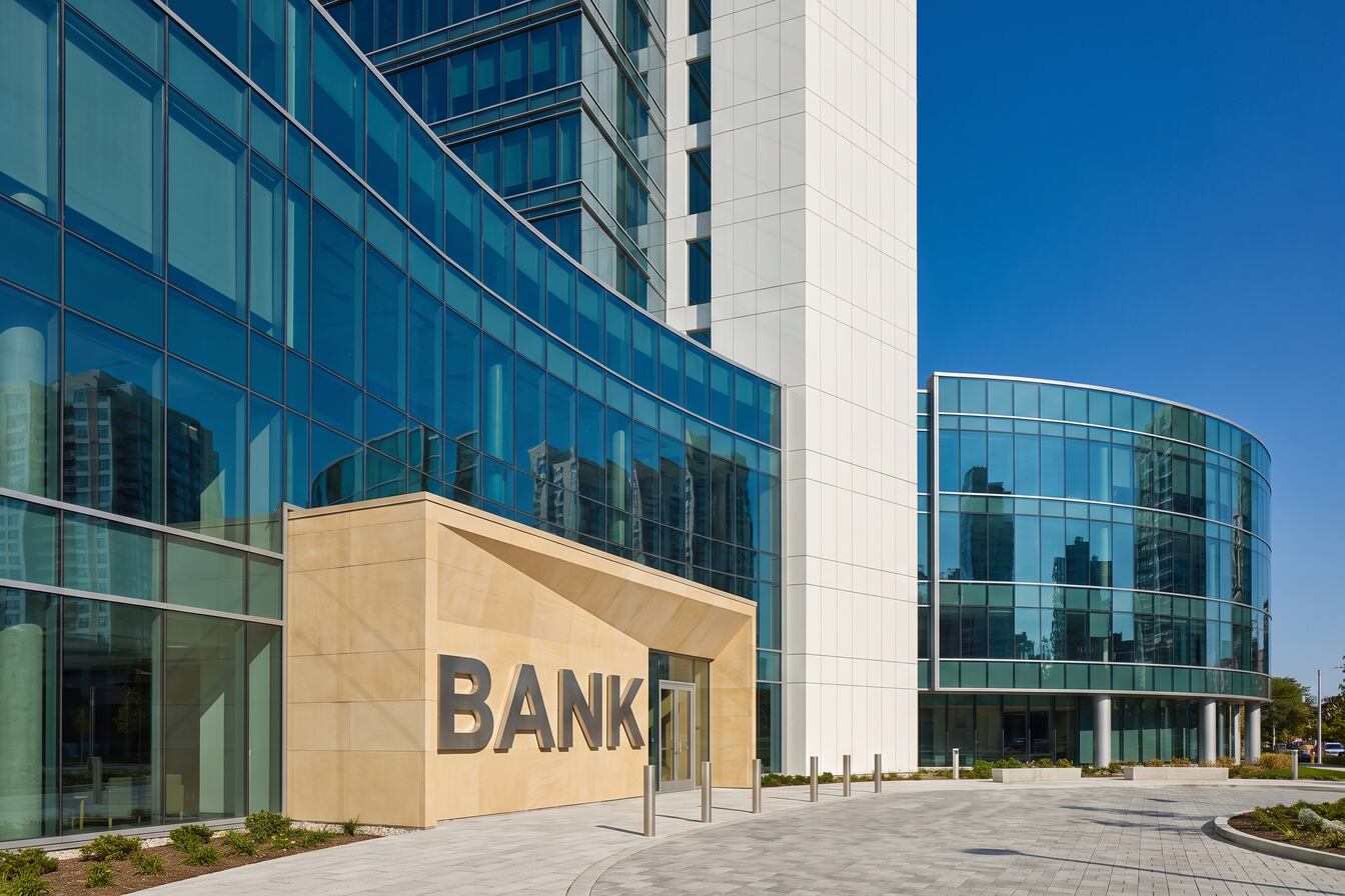 Modern bank building with large glass windows reflecting nearby high-rise buildings and a clear blue sky, featuring a beige stone entrance with the word 'BANK' in large letters.