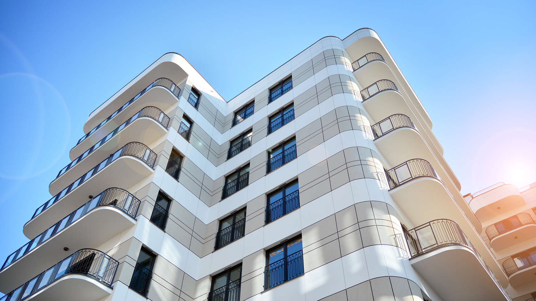 Low-angle view of a modern multi-story building with curved corners, balconies with black railings, and a blue sky background with sunlight flare.