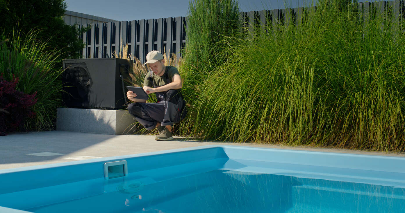 Technician wearing a cap and work uniform squatting beside a swimming pool, using a tablet near pool equipment and tall green grass.